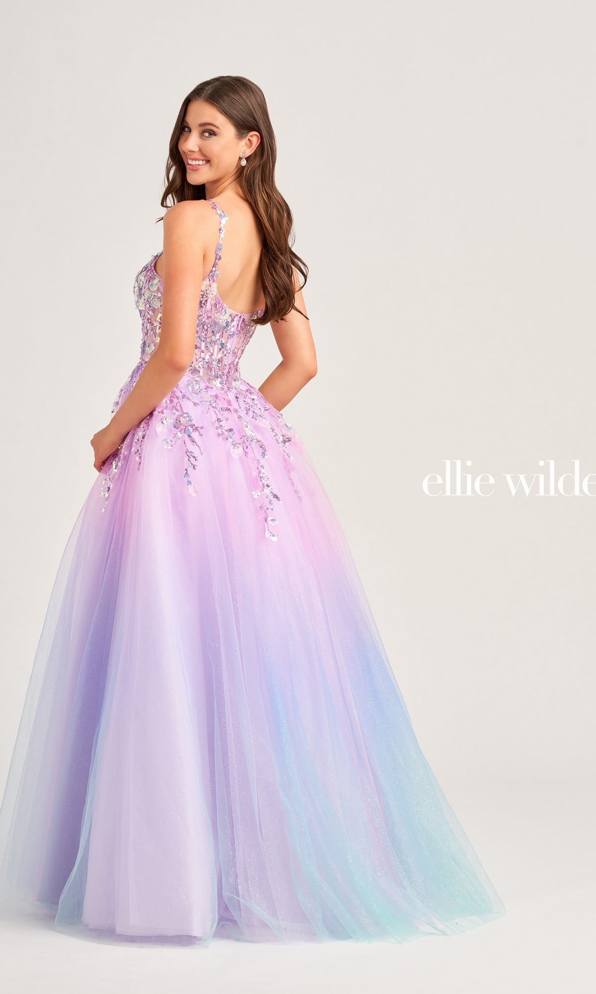 A woman in the Ellie Wilde Sequin Ombre Prom Ball Gown EW35055, featuring a fitted, beaded bodice and floor-length tulle skirt, stands facing away and smiles over her shoulder.